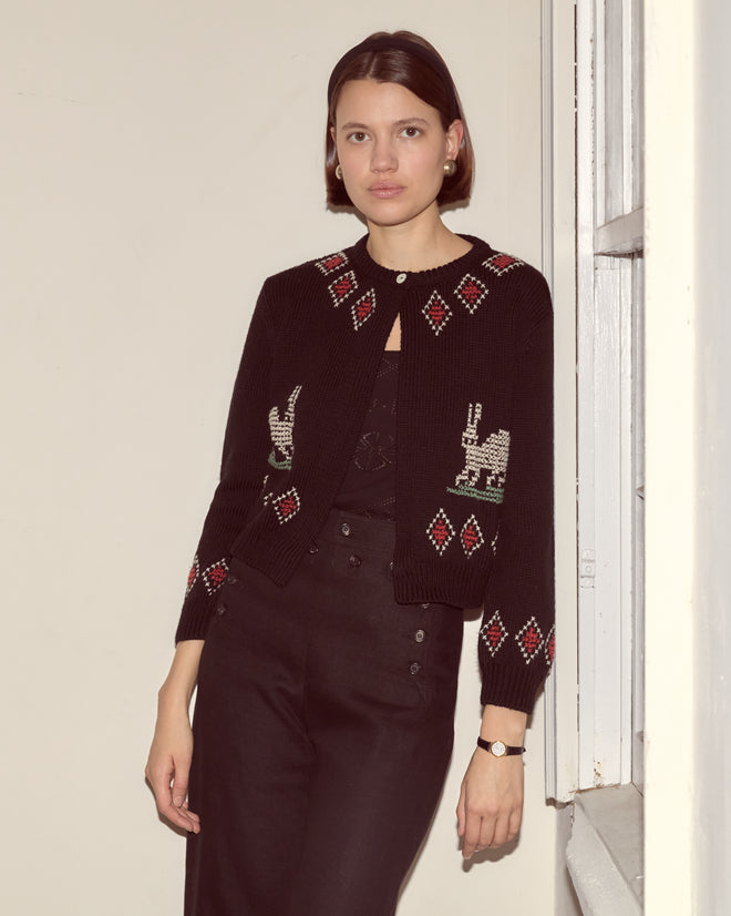 Woman wearing the Bunny Cardigan, a 100% wool knit with red and white patterns, stands by a window in softly lit surroundings.
