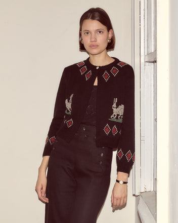 Woman wearing the Bunny Cardigan, a 100% wool knit with red and white patterns, stands by a window in softly lit surroundings.