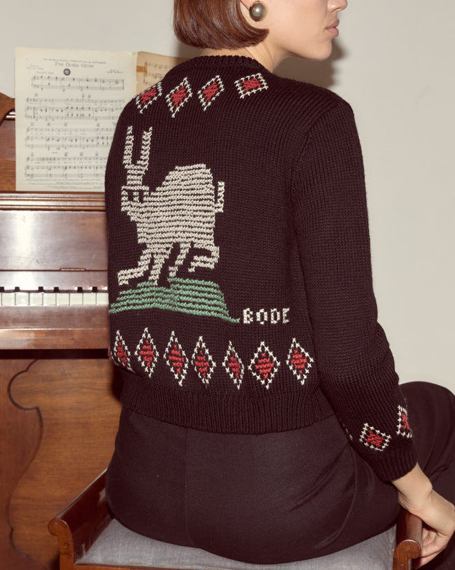 Woman in a Bunny Cardigan—a black knitwear sweater featuring a white bunny design—sits by a piano with sheet music on the stand.