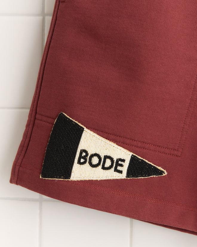 Close-up of Burgundy Pennant Shorts in 100% maroon cotton, featuring a black and white pennant patch with "BODE" lettering—evoking classic rugby shorts style.