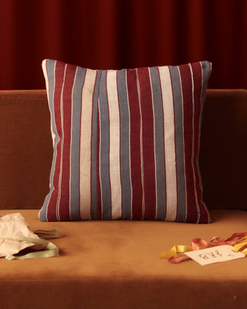 The Burgundy Multi-Stripe Large Pillow, handmade from deadstock strip-woven yardage in vibrant red, blue, and white, adds a touch of West African elegance to the brown sofa.