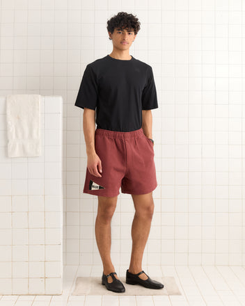 Young man in a black t-shirt and Burgundy Pennant Shorts stands in a white tiled room with a towel hanging on the wall.