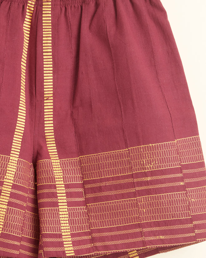 The Burgundy Railway Shorts - XXL, featuring vertical gold stripes and patterned trim, are crafted from strip-woven fabric and hang on a wooden hanger against a white wall.