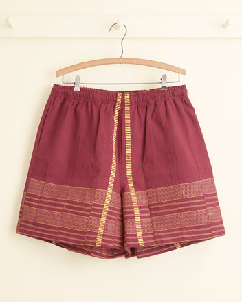 The Burgundy Railway Shorts - XXL, featuring vertical gold stripes and patterned trim, are crafted from strip-woven fabric and hang on a wooden hanger against a white wall.