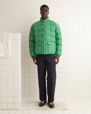 A person wearing a Burlington Puffer Jacket in green, which has a vintage-inspired style, along with dark pants stands in a tiled room facing forward.