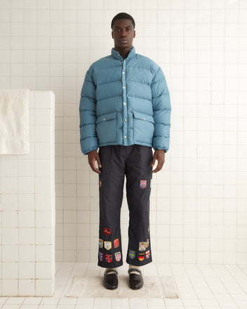 A man wearing the Burlington Puffer Jacket in blue and patch-decorated black pants, reminiscent of Olympic uniforms, stands on a white tiled floor.