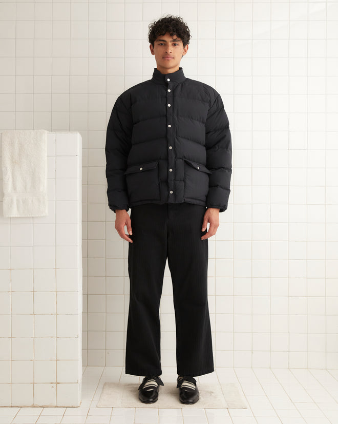 A person wearing a Burlington Puffer Jacket in black and pants stands in a tiled room, evoking the feel of vintage jackets. Nearby, a towel rests on a ledge, as if waiting for its moment.