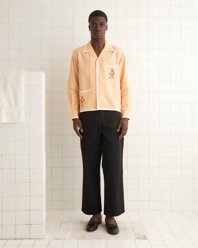 A man stands in a white-tiled room, wearing the Beaded Blackjack Long Sleeve Shirt in peach, which features a boxy fit and is made of a linen-cotton blend, paired with black pants. A towel hangs on the left wall.