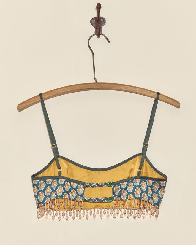 The Beaded Bouquet Bra features unique yellow and teal floral patterns, crafted from deadstock fabric with beaded fringe details, shown on a wooden hanger against a cream wall.