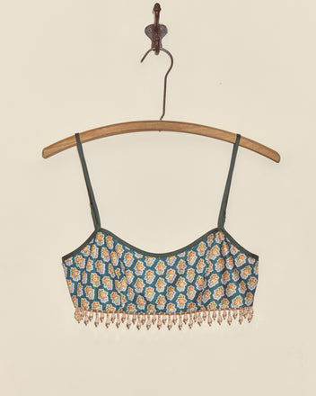 The Beaded Bouquet Bra, a green floral bralette crop top with thin straps and beaded fringe made from deadstock fabric, hangs on a wooden hanger against a white wall.
