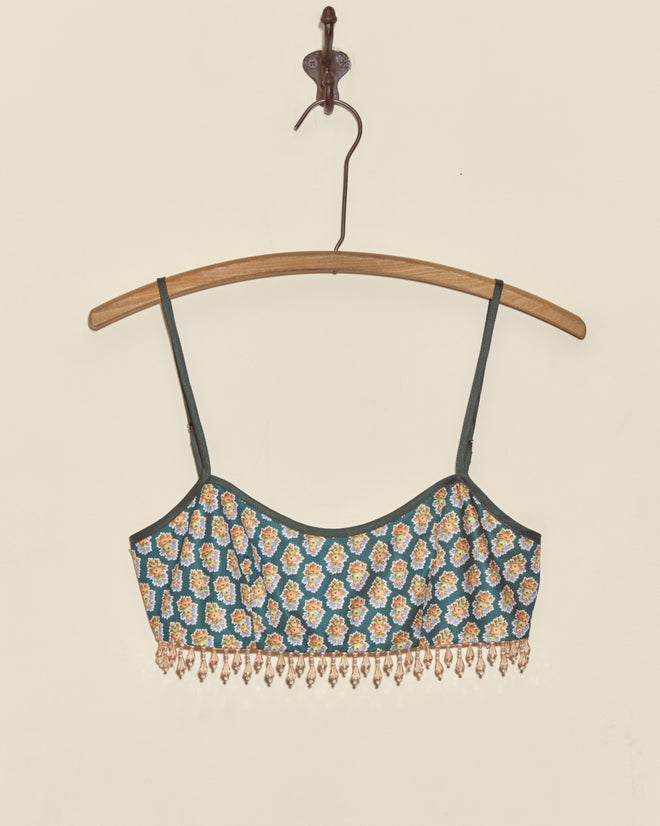 The Beaded Bouquet Bra, a green floral bralette crop top with thin straps and beaded fringe made from deadstock fabric, hangs on a wooden hanger against a white wall.