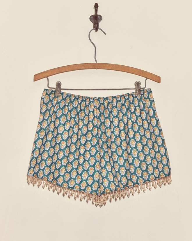 Beaded Bouquet Shorts in blue with a yellow floral bouquet pattern, crafted from deadstock fabric, hang on a wooden hanger against a cream wall.