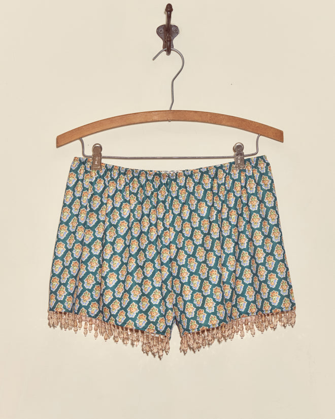 Beaded Bouquet Shorts with a yellow and white floral bouquet print and lace trim hang on a wooden hanger against a plain wall.