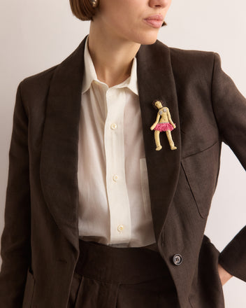 Woman in a brown suit and white shirt wears the Calder Fanni Brooch, inspired by Alexander Calder’s jewelry and shaped like a doll in a pink skirt, reminiscent of the Calder Circus brooch from the Whitney Museum collection.