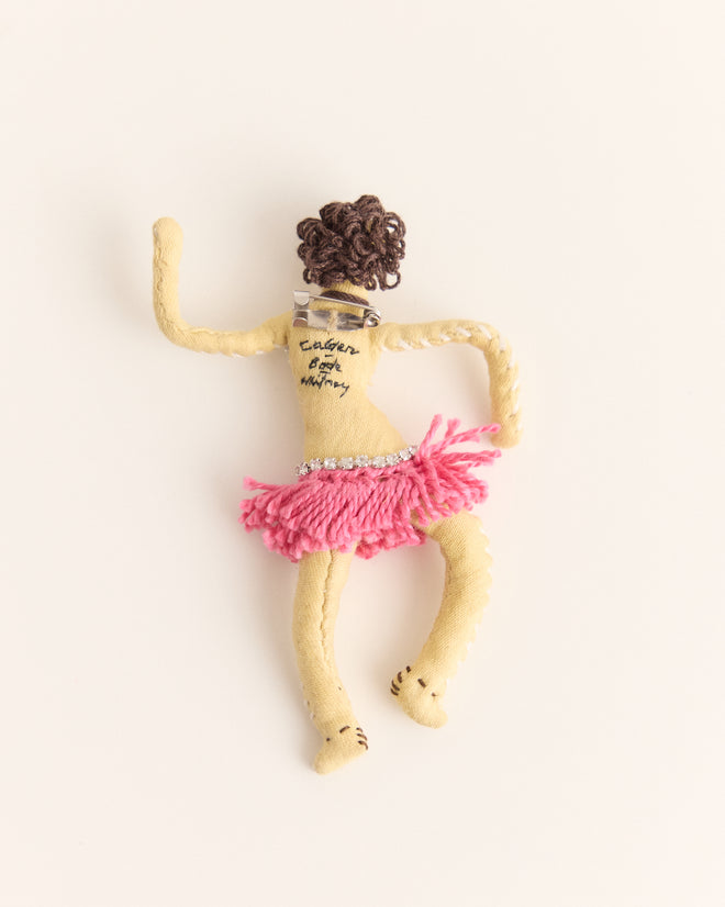 The Calder Fanni Brooch, inspired by the whimsical spirit of Alexander Calder jewelry, features a handmade doll with a pink skirt, curly hair, and back text, displayed on a plain background.