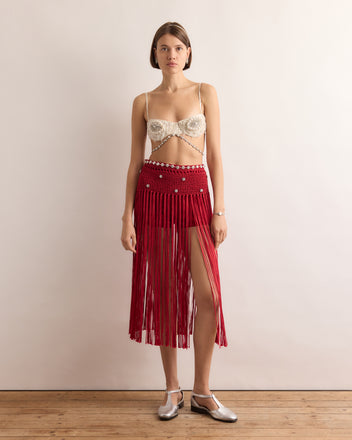 Woman in the Calder Fanni Fringe Skirt, featuring red fringe and a thigh slit, stands on a wooden floor against a plain wall—channeling the playful spirit of Calder’s Circus as seen at the Whitney Museum of American Art.