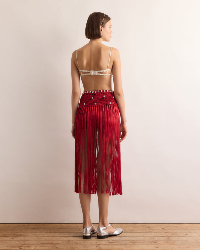 A woman stands facing away, wearing a beaded bra and the Calder Fanni Fringe Skirt with silver shoes on a wooden floor—her pose echoing the playful spirit of performers from Alexander Calder’s Circus, now at the Whitney Museum.