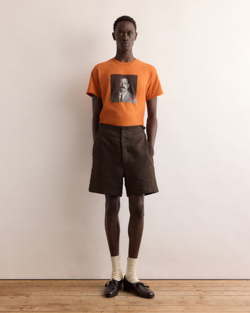 A person wears the Calder Identity Card Tee—an orange graphic T-shirt—paired with brown shorts, cream socks, and black loafers against a plain background.