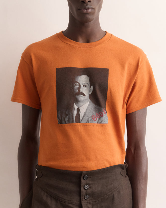 Person wearing the Calder Identity Card Tee, an orange t-shirt with a black-and-white portrait of Alexander Calder, the artist behind Calder’s Circus, printed on the front.