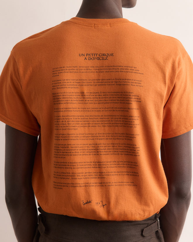 Person wearing the Calder Identity Card Tee, an orange t-shirt with French text on the back, inspired by the playful spirit of Alexander Calder’s Circus, facing away from the camera.