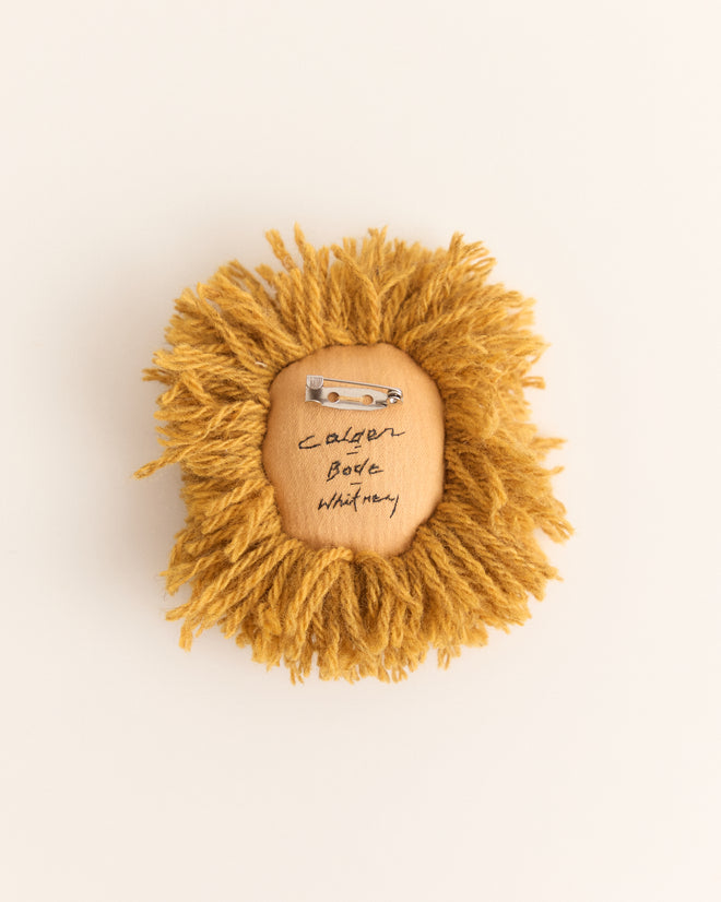 The Calder Leo Brooch is a round, golden-yellow yarn accessory with a pin back and handwritten text on its wooden center, inspired by Whitney Museum collaborations and reminiscent of Alexander Calder’s jewelry.
