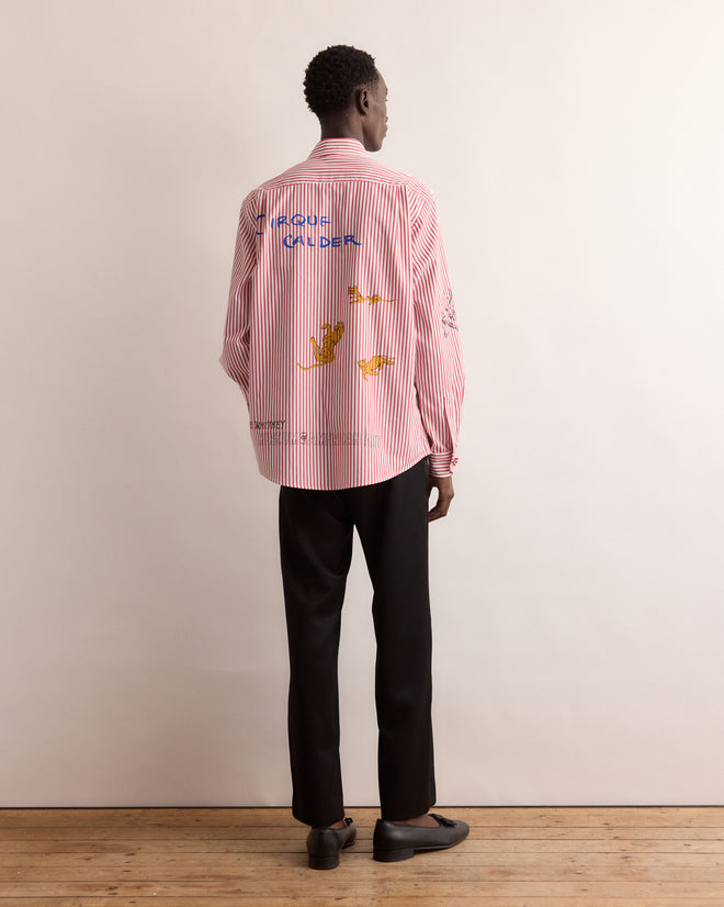 A person stands facing away, wearing the Calder Painted Cirque Long Sleeve Shirt with hand-painted red stripes and colorful circus-inspired drawings, paired with black pants.