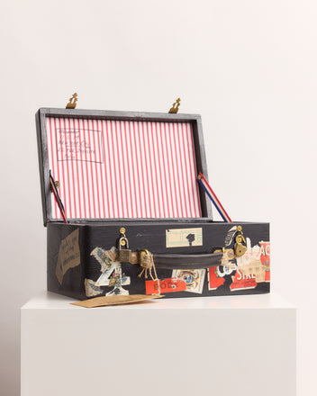 The Calder Suitcase Box, inspired by Calder’s Circus and showcased by the Whitney Museum of American Art, sits open on a white pedestal, its travel stickers and red striped interior evoking a vintage, playful charm.