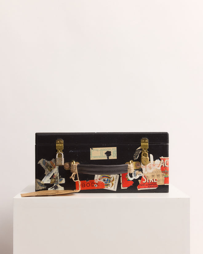 The Calder Suitcase Box, a black vintage-inspired piece adorned with travel stickers, is displayed on a white pedestal against a plain background.