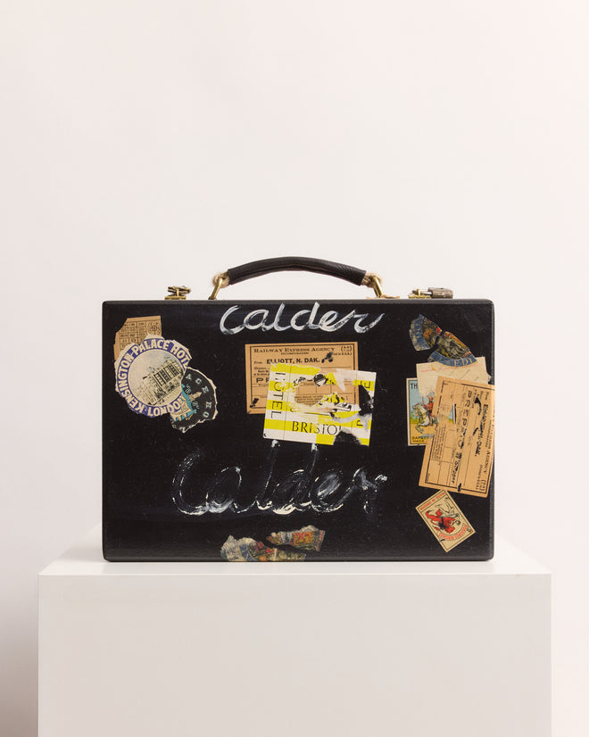 The Calder Suitcase Box, a black case adorned with vintage travel stickers and "Calder" lettering, is displayed on a white pedestal and recalls Calder’s Circus, now preserved by the Calder Foundation.