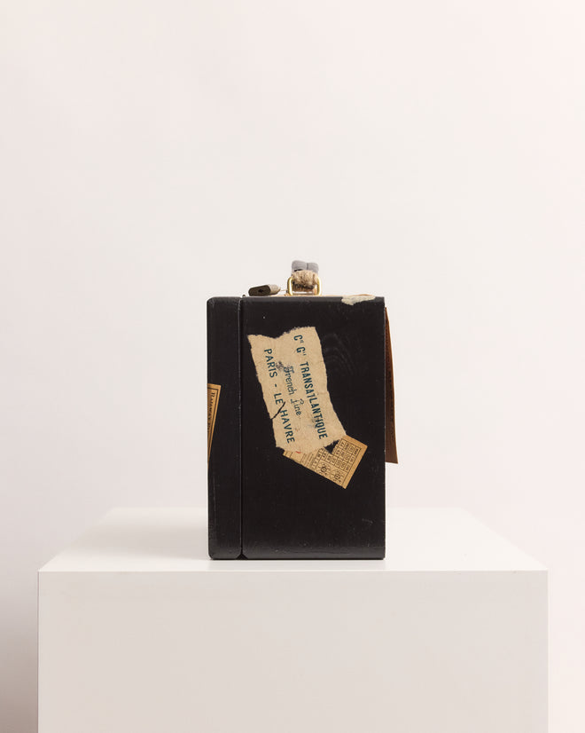 The Calder Suitcase Box, with its vintage black design and worn label, sits on a white pedestal against a plain background, evoking displays at the Whitney Museum of American Art and reminiscent of Calder’s Circus.