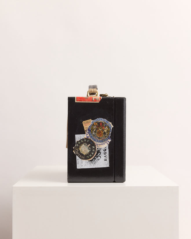 The Calder Suitcase Box, a black suitcase adorned with travel stickers, stands upright on a white pedestal against a plain background, evoking the spirit of Calder’s Circus exhibited at the Whitney Museum of American Art.