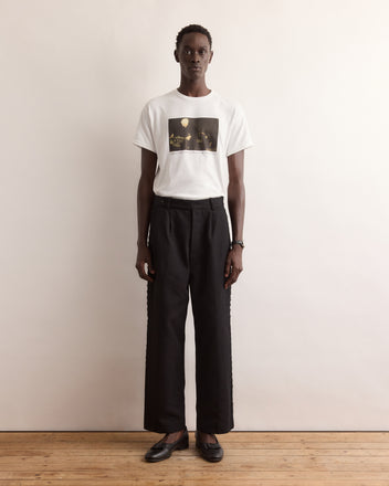 A person stands against a wall wearing the Calder Suitcase Tee, a white graphic T-shirt inspired by Alexander Calder’s Circus, paired with black pants and black shoes.