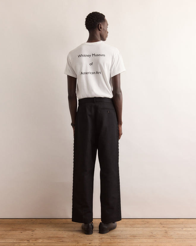 A person stands on a wooden floor wearing black pants and the Calder Suitcase Tee, a white T-shirt featuring "Whitney Museum of American Art" on the back, inspired by Alexander Calder’s iconic Circus.