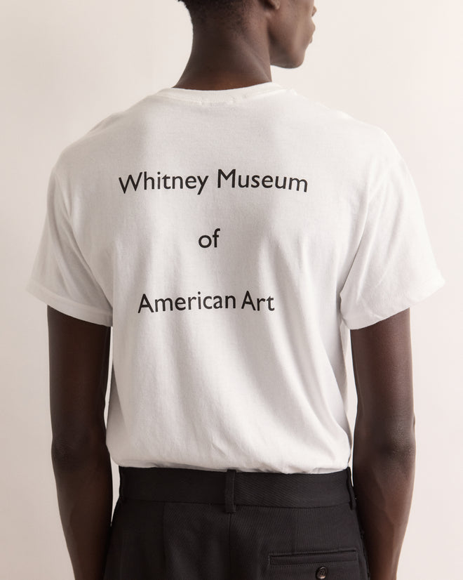 A person faces away wearing the Calder Suitcase Tee—a white shirt featuring “Whitney Museum of American Art” on the back, inspired by exhibitions like Alexander Calder’s Circus at the Whitney.