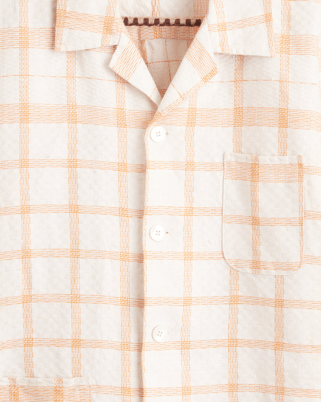 A Callaway Peach Shirt in M/L, featuring a short-sleeve button-up design with a pale orange plaid pattern, hangs on a hook. It boasts two unique pockets on the left side of the chest and near the hem, making it truly one-of-a-kind.