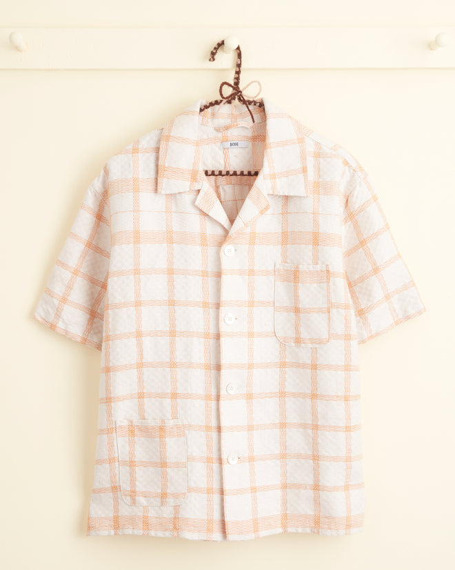 A Callaway Peach Shirt in M/L, featuring a short-sleeve button-up design with a pale orange plaid pattern, hangs on a hook. It boasts two unique pockets on the left side of the chest and near the hem, making it truly one-of-a-kind.