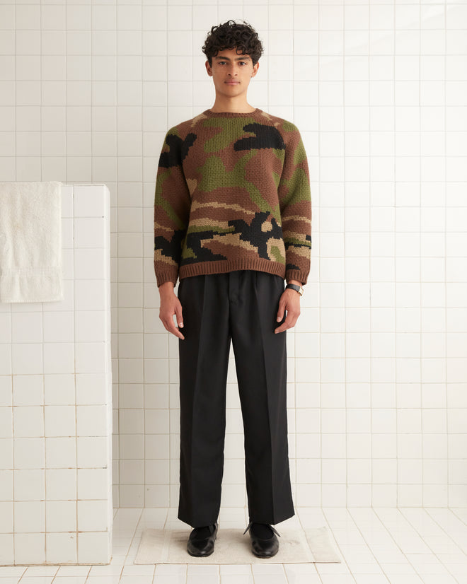 A person wearing a Camouflage Crewneck sweater and black pants stands in a tiled room.