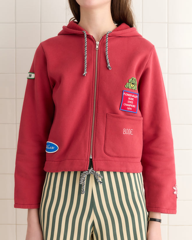 A person in a Camper Hoodie, featuring a red cotton fleece design with vintage patches, paired with green striped pants stands against a tiled background.