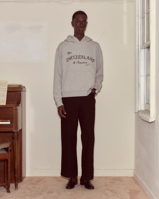 A person wearing the Campground Hoodie, featuring a log cabin illustration and advertising slogan, with black pants stands indoors by a piano and a window.