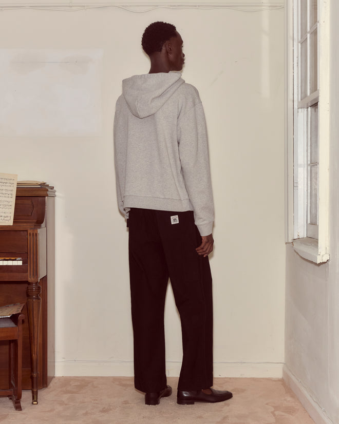 Wearing a Campground Hoodie made of 100% cotton and black pants, a person stands facing a wall near a piano and window at Camp Geneva.