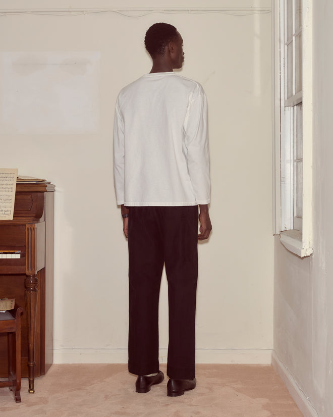 A person stands facing a plain wall, wearing a white Campground Long Sleeve shirt and black pants.