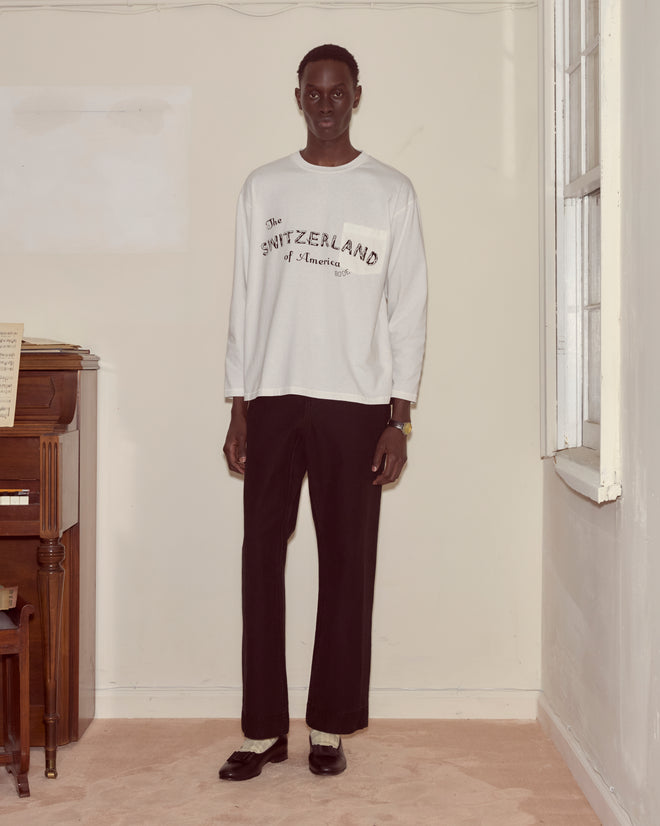 A person in a white Campground Long Sleeve and black pants stands indoors by a piano and window, their outfit featuring the classic 100% cotton camp tee.