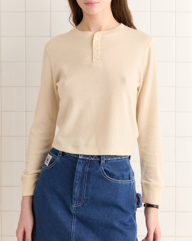 An individual wearing a Campus Henley in beige, made from organic cotton, paired with a denim skirt stands against a tiled wall.