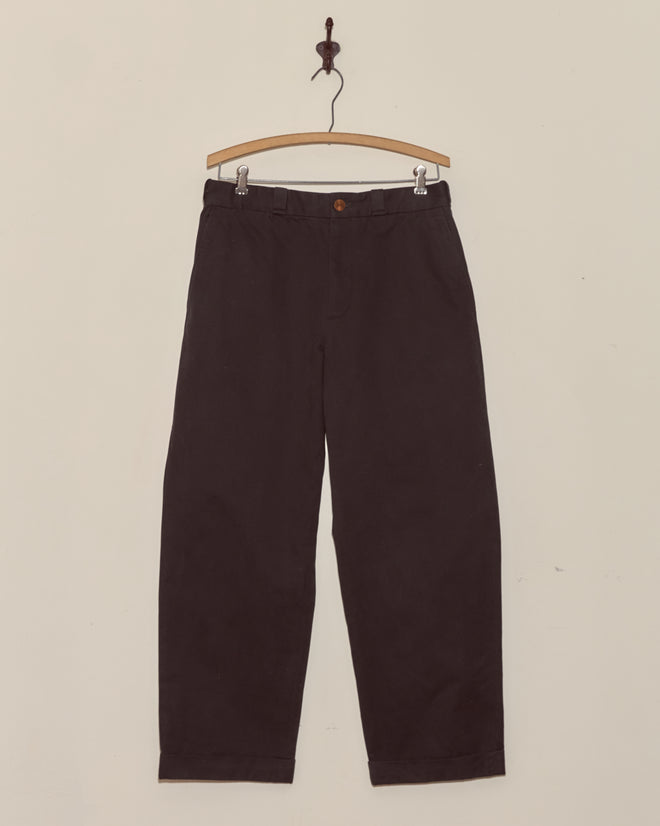 A pair of Campus Trousers - Black in heavyweight cotton twill hangs on a wooden hanger against a plain white wall.