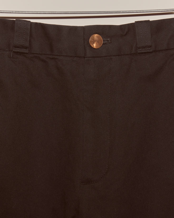 Close-up of the waistband and button on the Campus Trousers - Black, made from heavyweight cotton twill, displayed on a metal clothing rack.