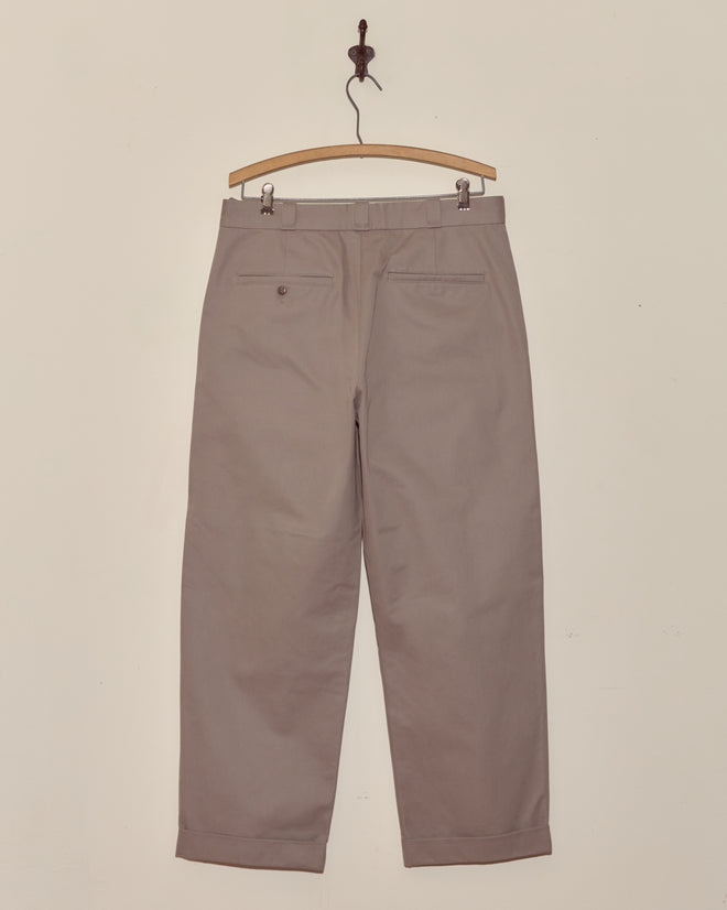 A pair of Campus Trousers - Grey in heavyweight cotton twill hangs on a wooden hanger against a plain light-colored wall.