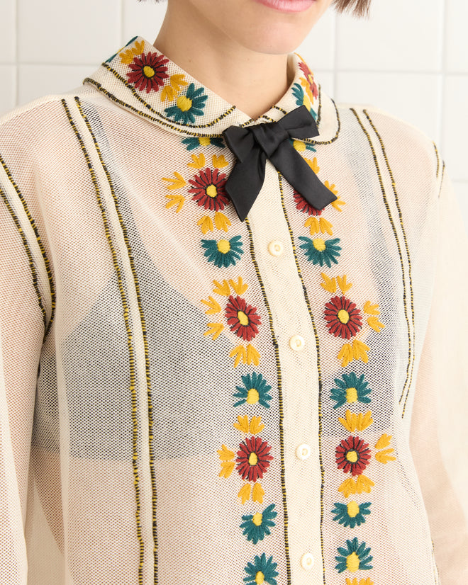 A close-up of a person wearing the Candyfloss Blouse, a sheer cotton top with colorful floral embroidery, a black ribbon bow, and an elegant organza collar.