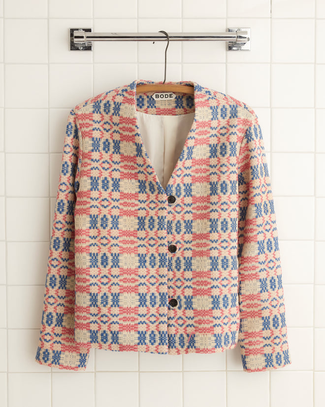 The Candyfloss Coverlet Jacket - 2, featuring a red, blue, and beige geometric design reminiscent of an antique coverlet, hangs elegantly on a tiled wall.