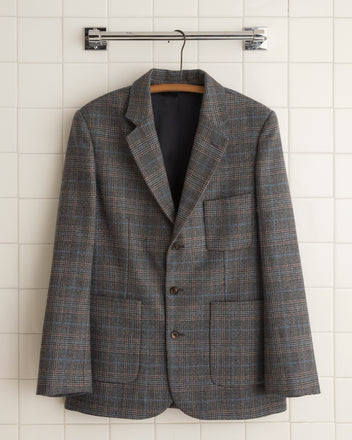 The Cannery Plaid Suit Jacket - 44, crafted from fine suiting fabric, hangs elegantly on a wooden hanger against a white tiled wall.