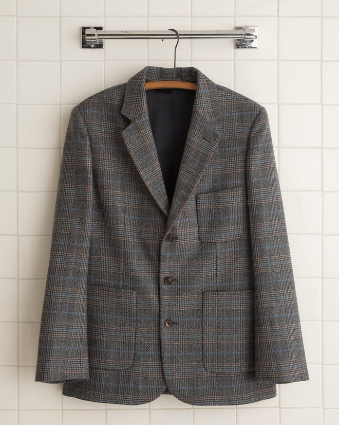 The Cannery Plaid Suit Jacket - 44, crafted from fine suiting fabric, hangs elegantly on a wooden hanger against a white tiled wall.